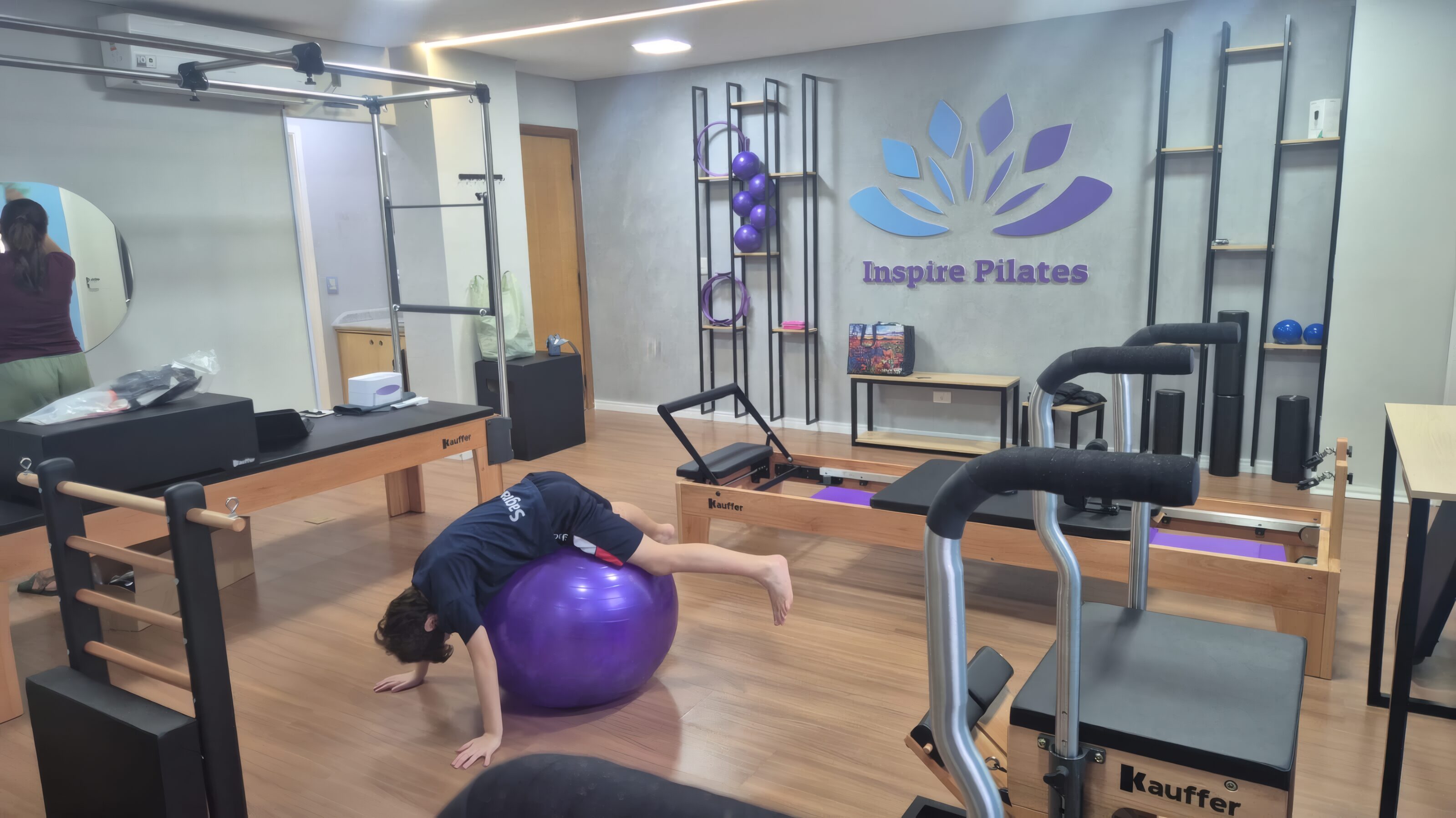 Salão do Inspire Pilates: aparelhos de madeira clara, bola roxa e a parede com o logo do estúdio