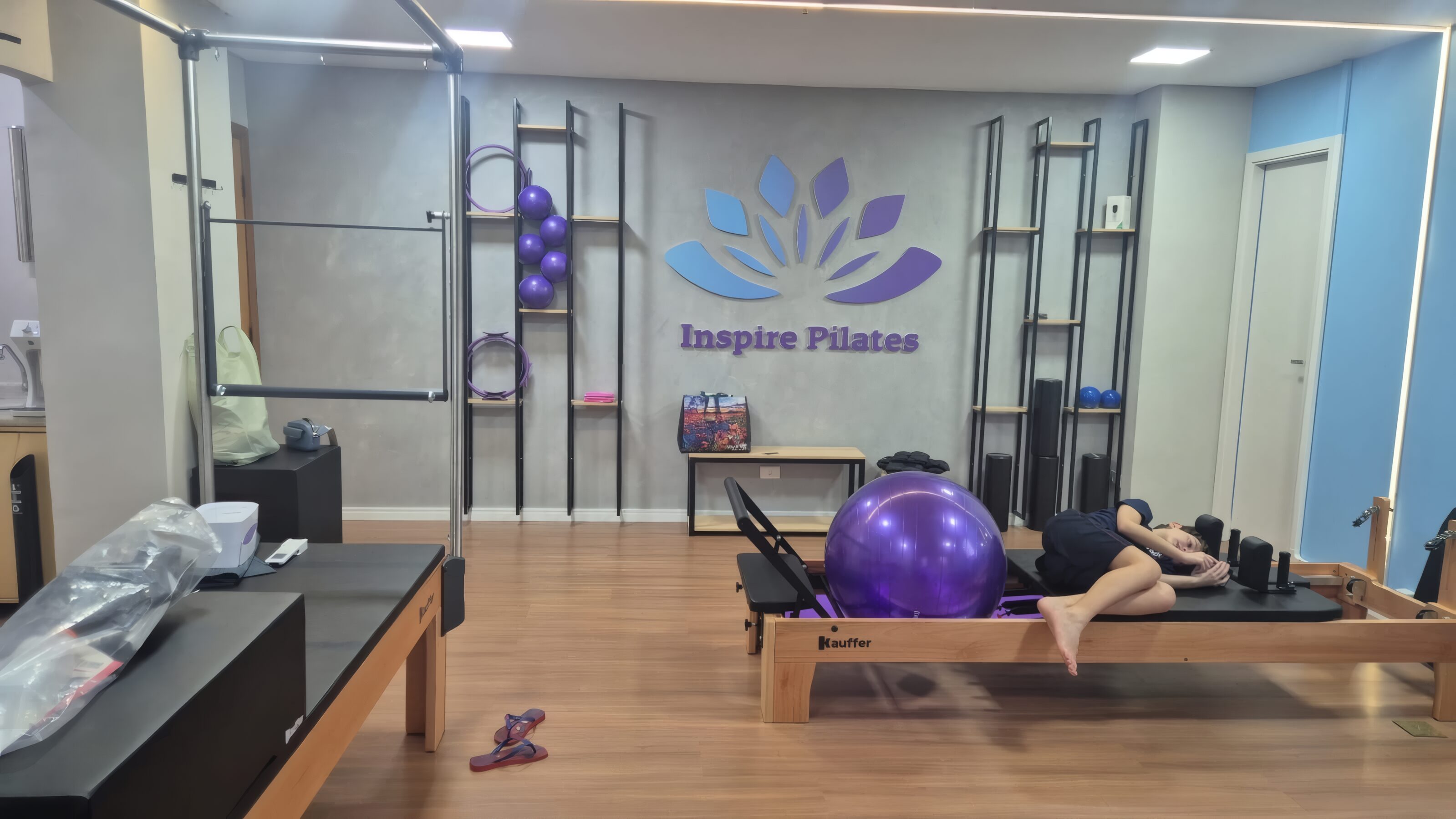 Vista geral do estúdio, com Reformer, bola de pilates e a parede de concreto com o logo